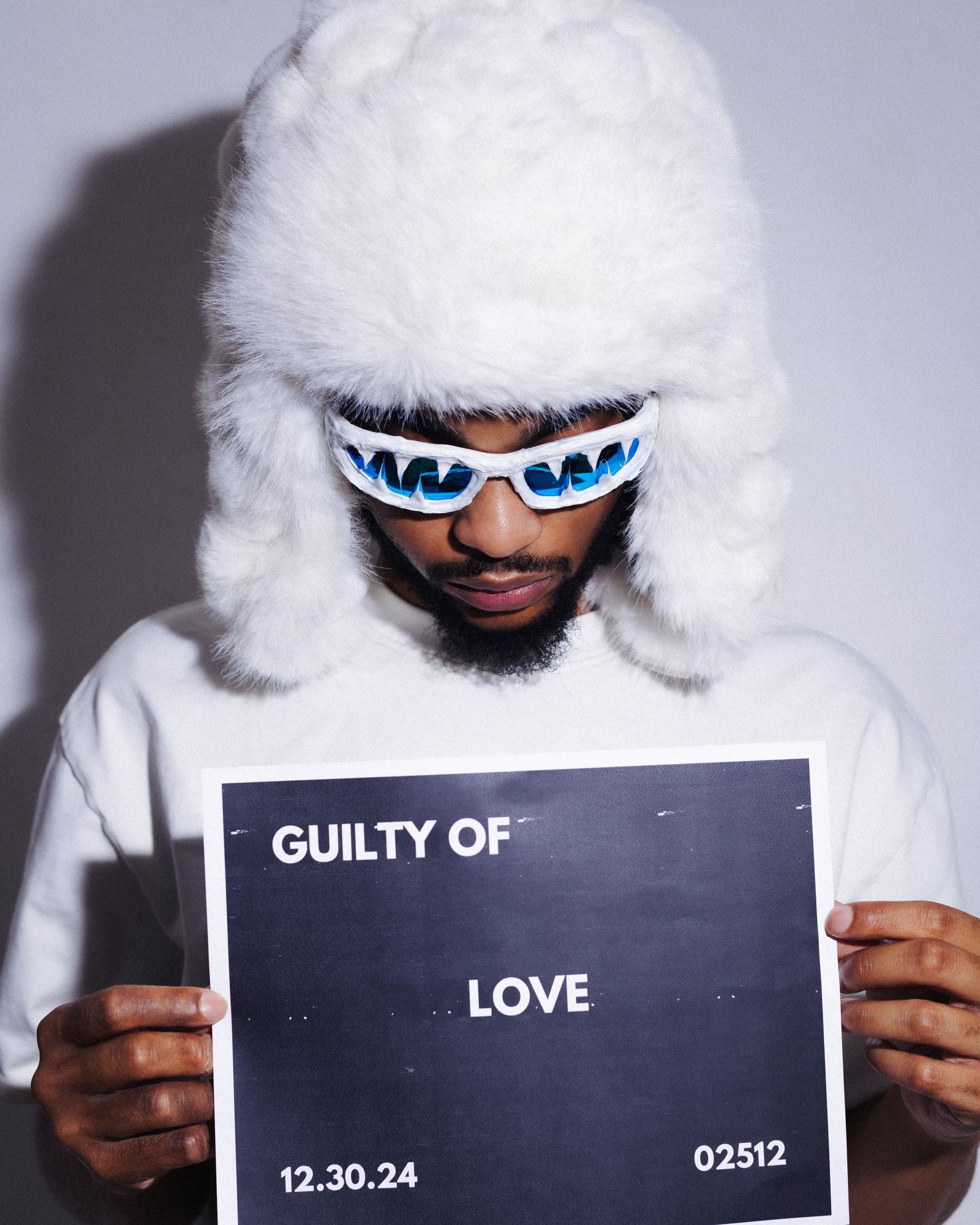 Self-portrait in a tall white fur hat and white wraparound sunglasses, holding a printed sign that reads "Guilty of Love, 12.30.24".