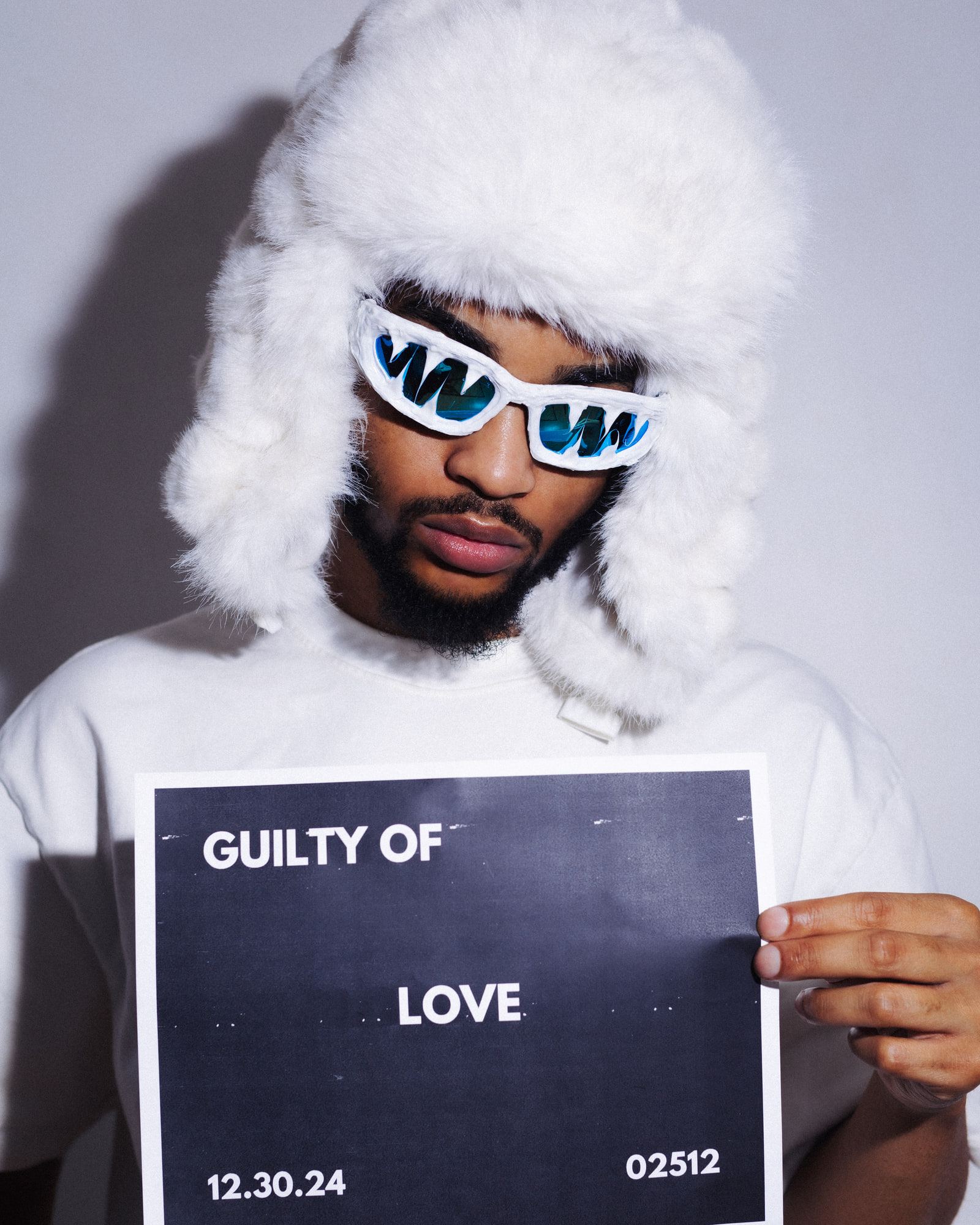 Self-portrait in the white fur hat and sunglasses, looking down at the camera, holding the "Guilty of Love" sign.