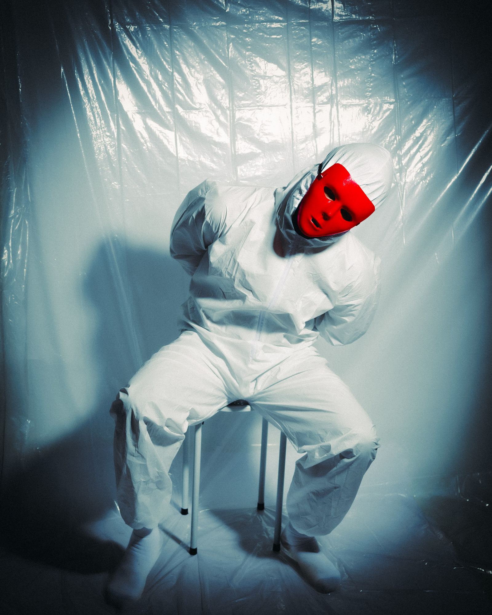 Self-portrait in a white hazmat suit and a glossy red face mask, seated on a stool in front of a plastic sheet lit from behind.