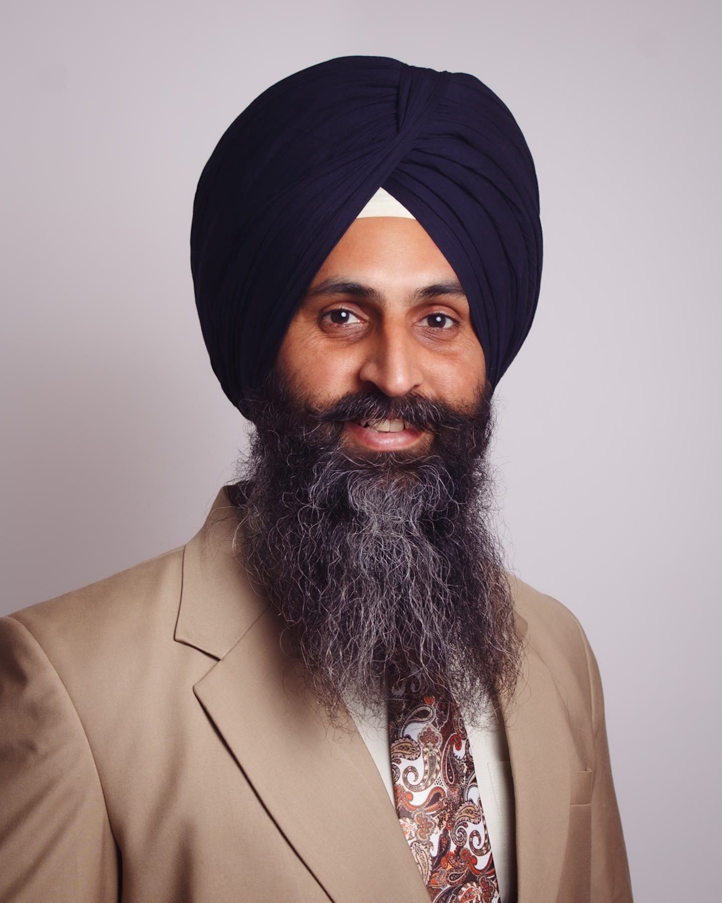 Portrait of a smiling man with a long grey beard in a navy turban, beige suit and patterned tie.