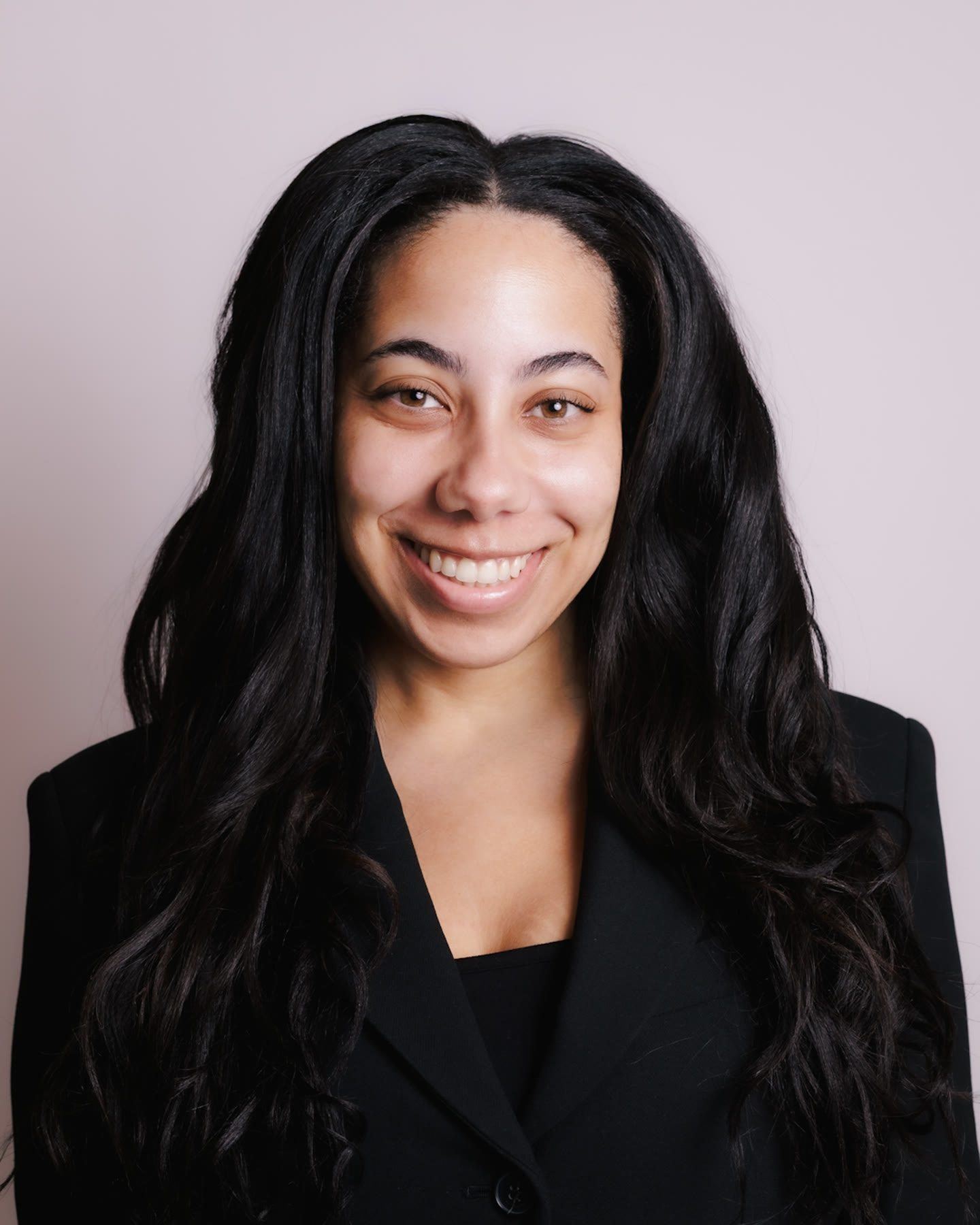 Headshot of a smiling woman with long dark curly hair in a black blazer, against a soft pink backdrop.