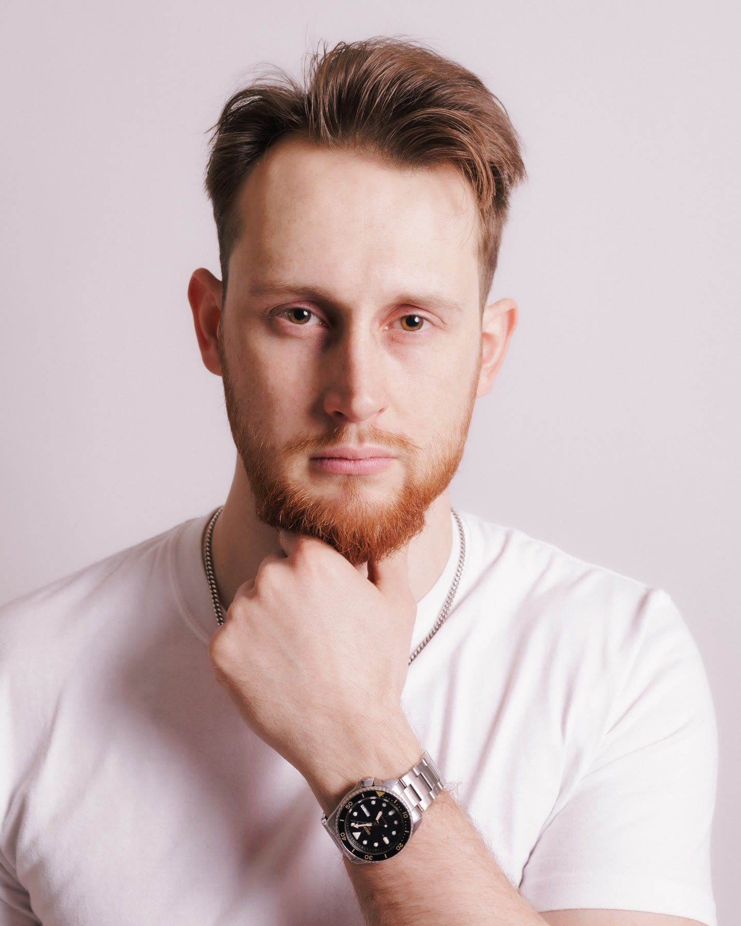 Headshot of a man with ginger hair and beard in a white T-shirt, his hand at his chin.