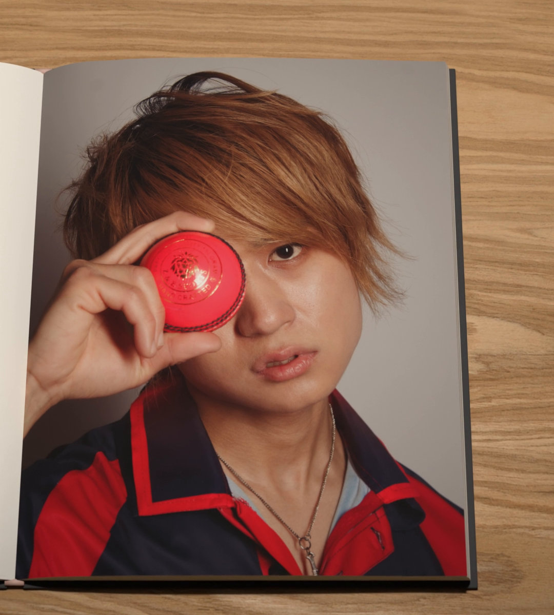 A person with copper hair and a red track jacket holds a red ball over one eye.