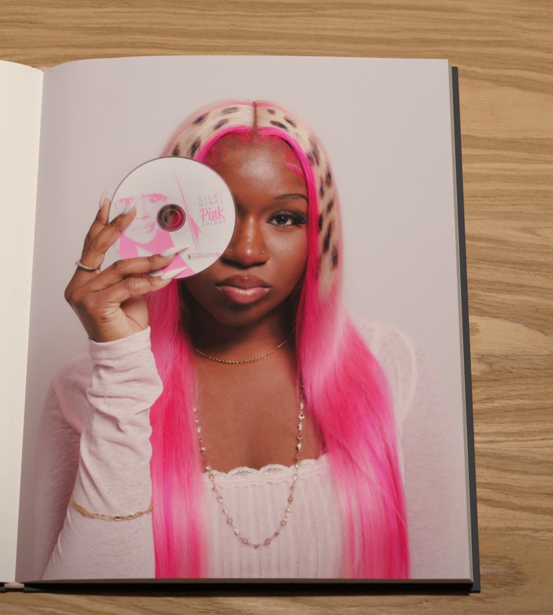 A person with long pink hair holds a CD over one eye.