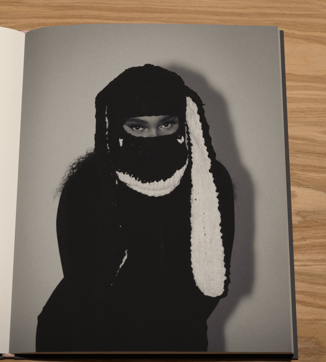 In black and white, a person in a black knitted hood with long white knitted ears, the face covered below the eyes.