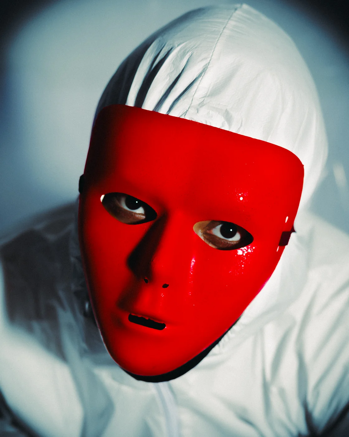 Self-portrait in a white hooded suit and a glossy red mask, one eye looking out.