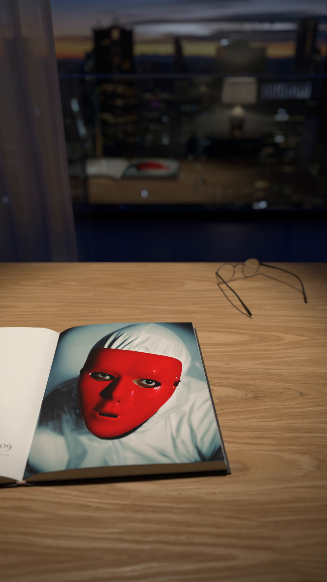 A magazine open on a desk at dusk, showing a portrait of a person in a red mask, with a pair of glasses beside it and a city skyline through the window.