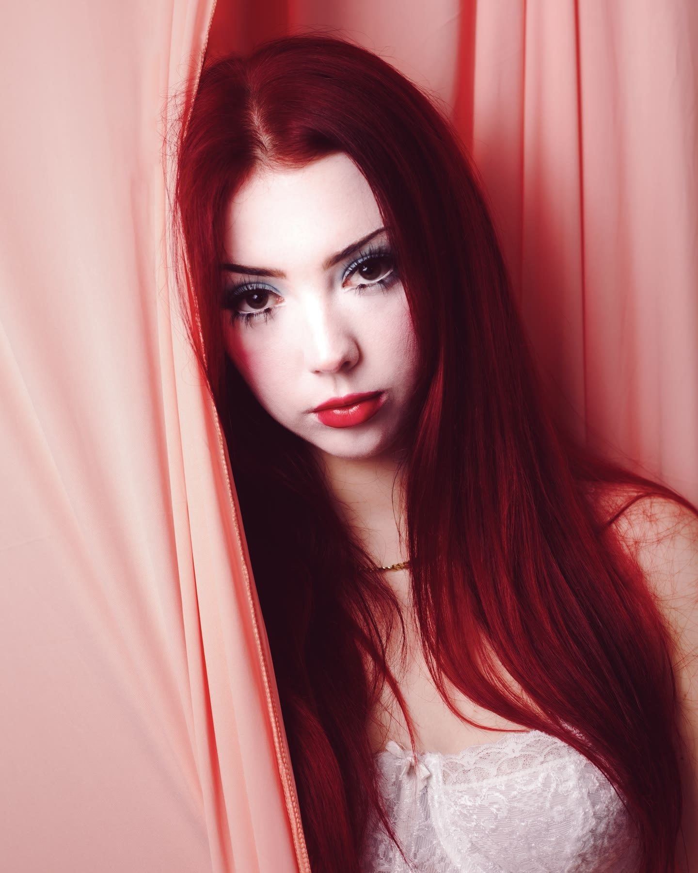 A woman with long red hair half hidden behind a pink curtain, looking at the camera.