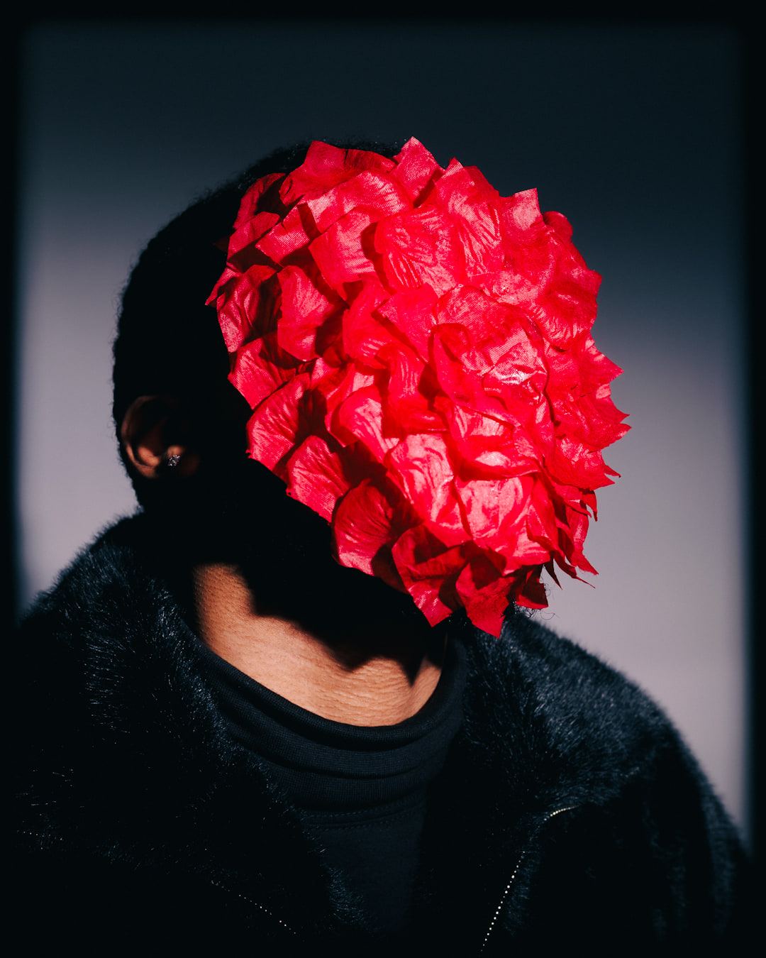 Self-portrait in a black fur jacket, face hidden behind a dense mask of red petals, lit against a grey wall.