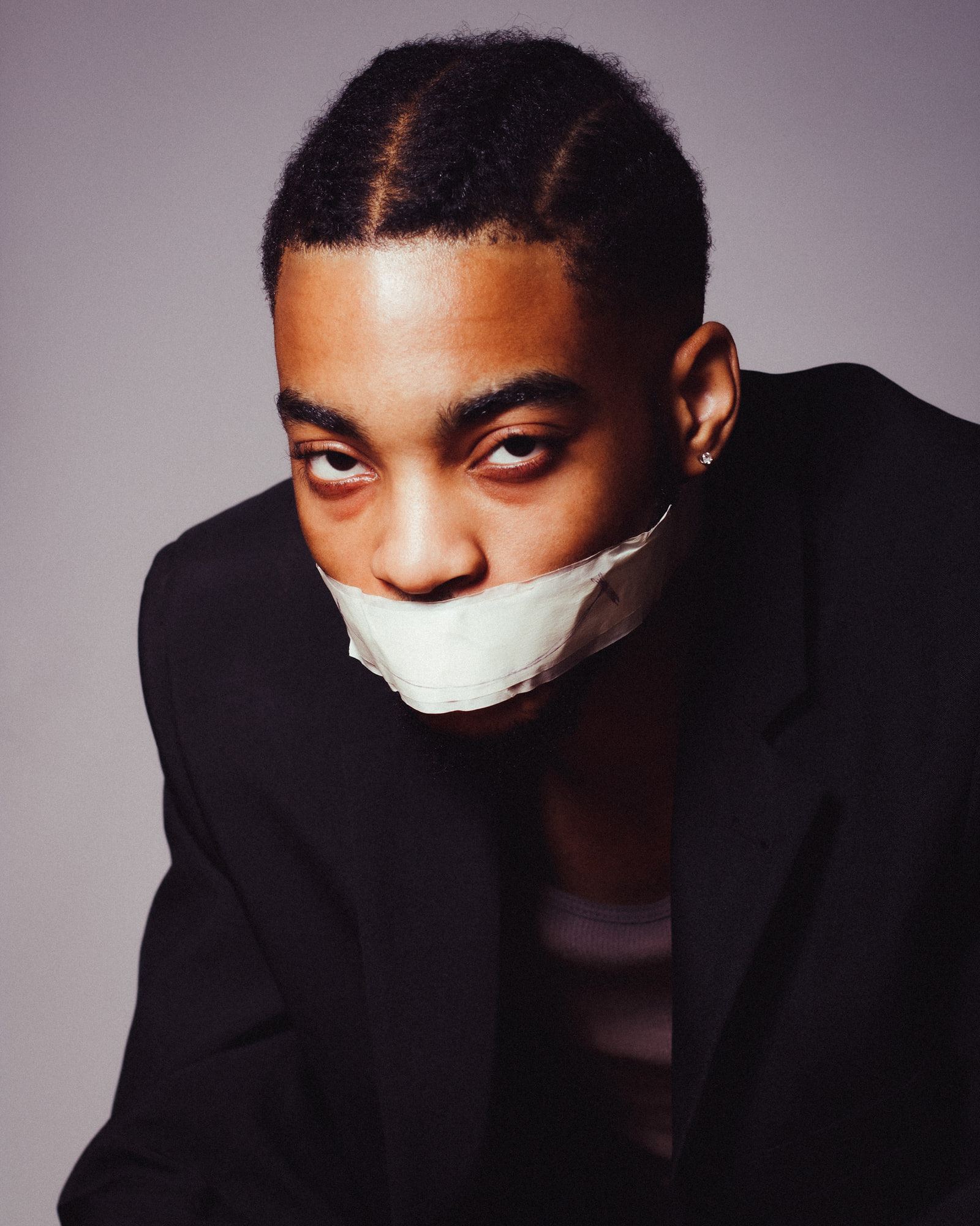 Self-portrait in a black blazer with a strip of white tape over the mouth, looking straight at the camera.