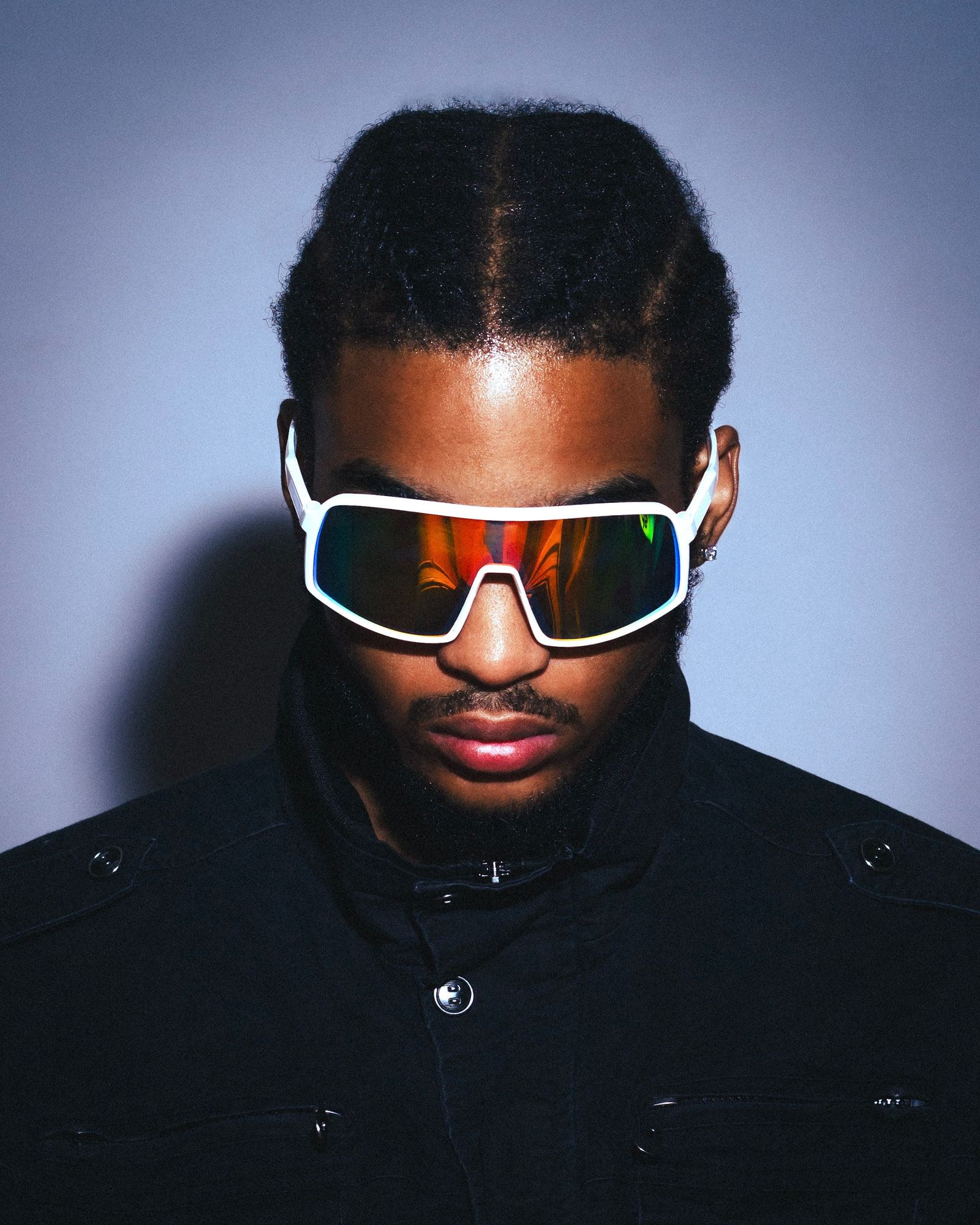 Self-portrait looking down, wearing white-framed shield sunglasses with orange lenses and a black jacket, against a pale blue wall.